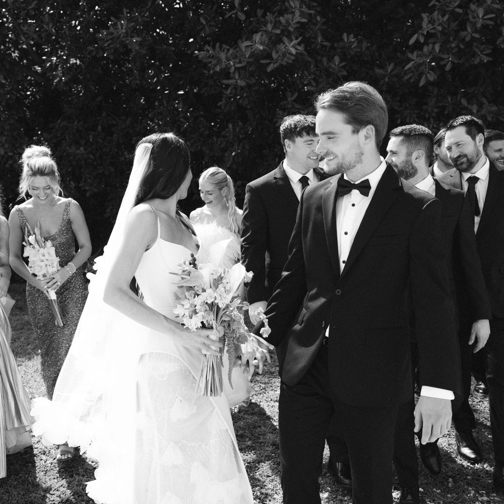 A black and white wedding photo showing a joyful moment between newlyweds and their wedding party outdoors.