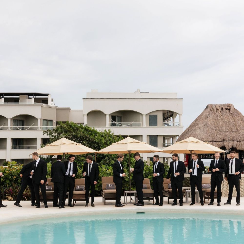 Wedding party in dark suits standing in line by a resort pool with beige umbrellas and thatched roof building in background.