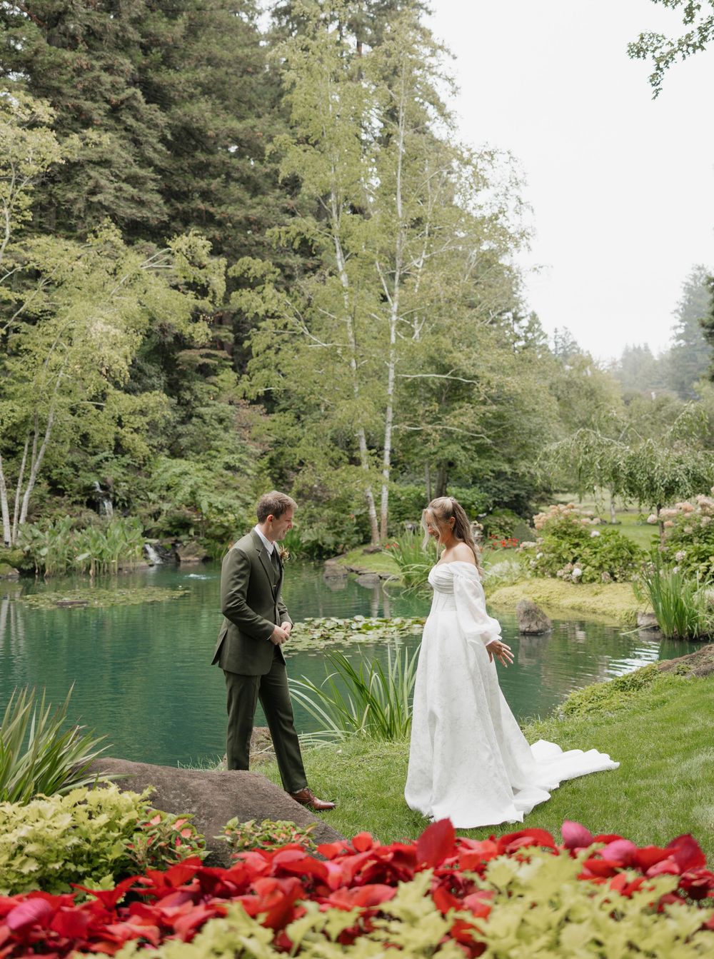 A couple stands by a serene turquoise pond surrounded by evergreen trees and red poinsettias in a garden setting.
