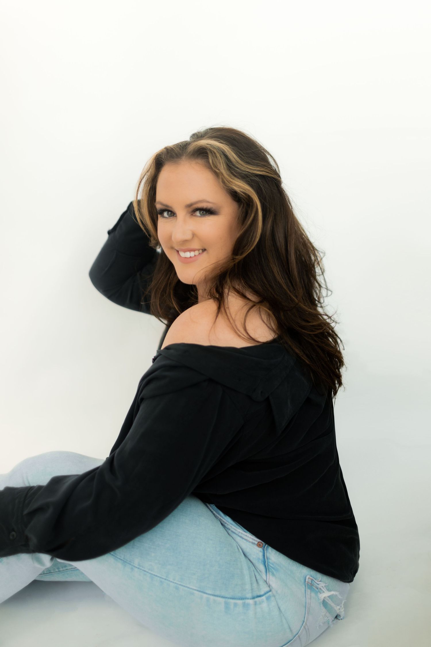 Portrait of someone wearing a black off-shoulder sweater and light jeans, posing against a white background.