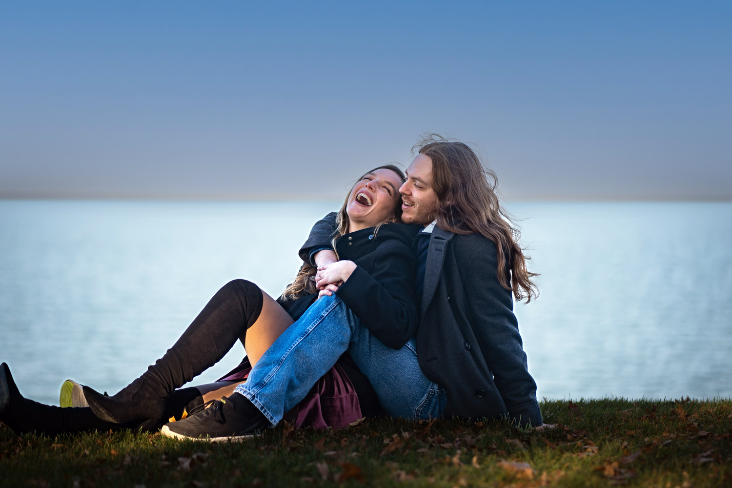 Two people sit close together outdoors at dusk sharing a laugh while resting against a white wall.