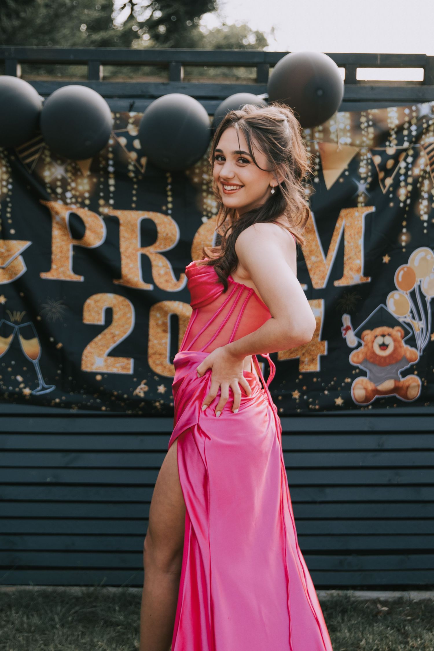 A student in a bright pink formal gown poses in front of a prom backdrop with black balloons and decorations.