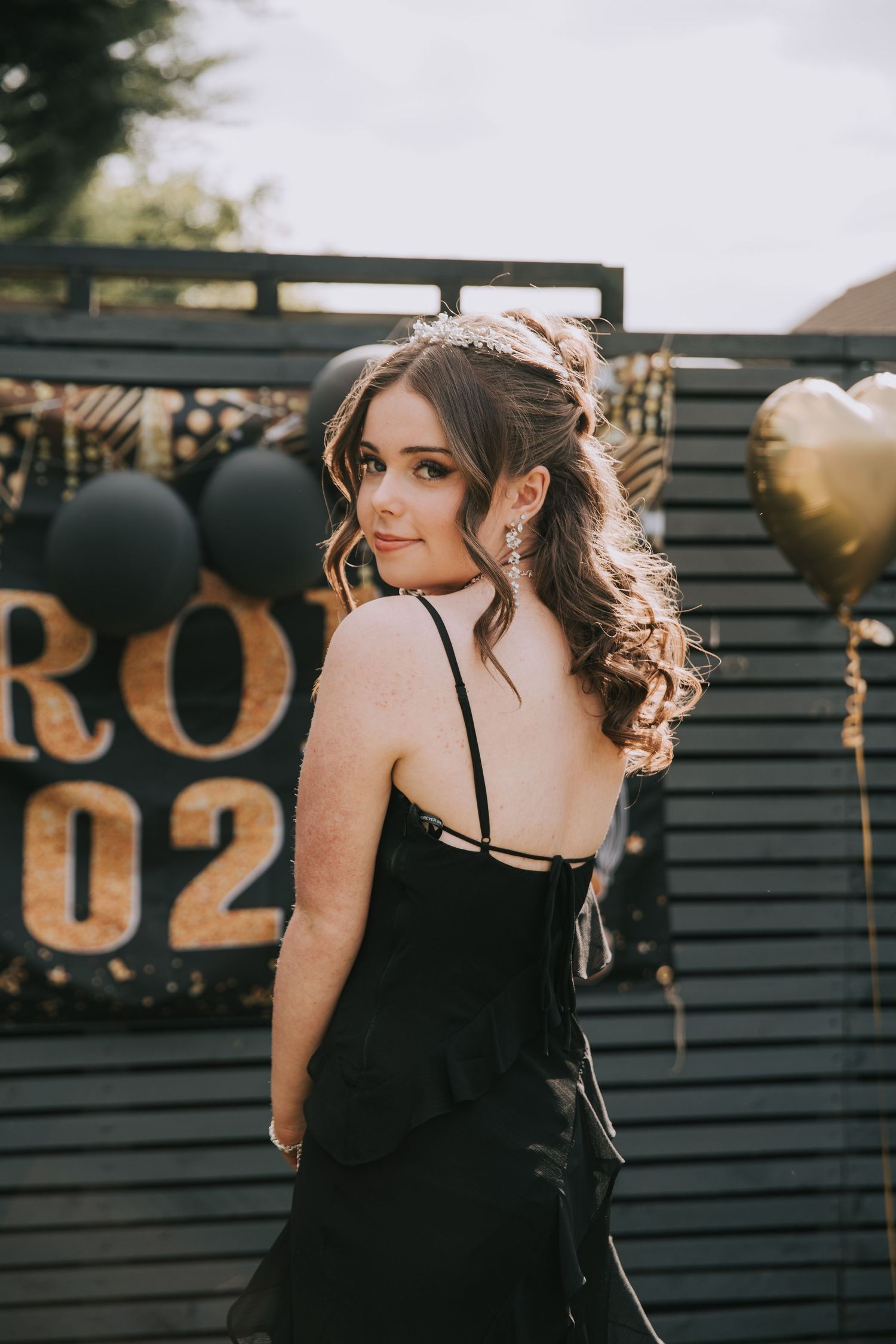 A woman in an elegant black dress poses at a prom event with gold and black balloon decorations in the background.