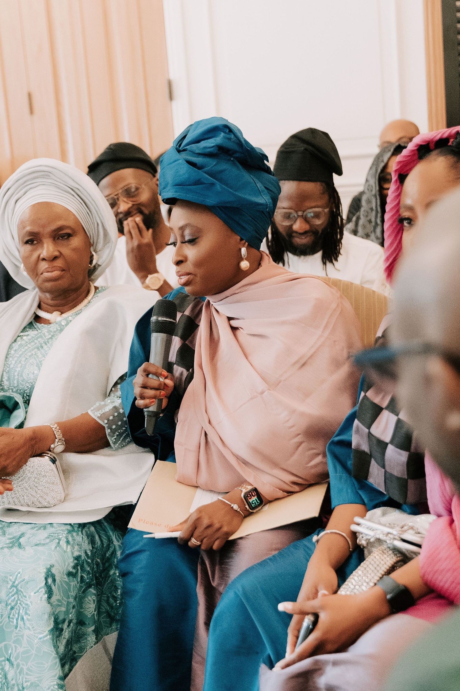 A group in traditional African attire sits together at an event, with one person speaking into a microphone.