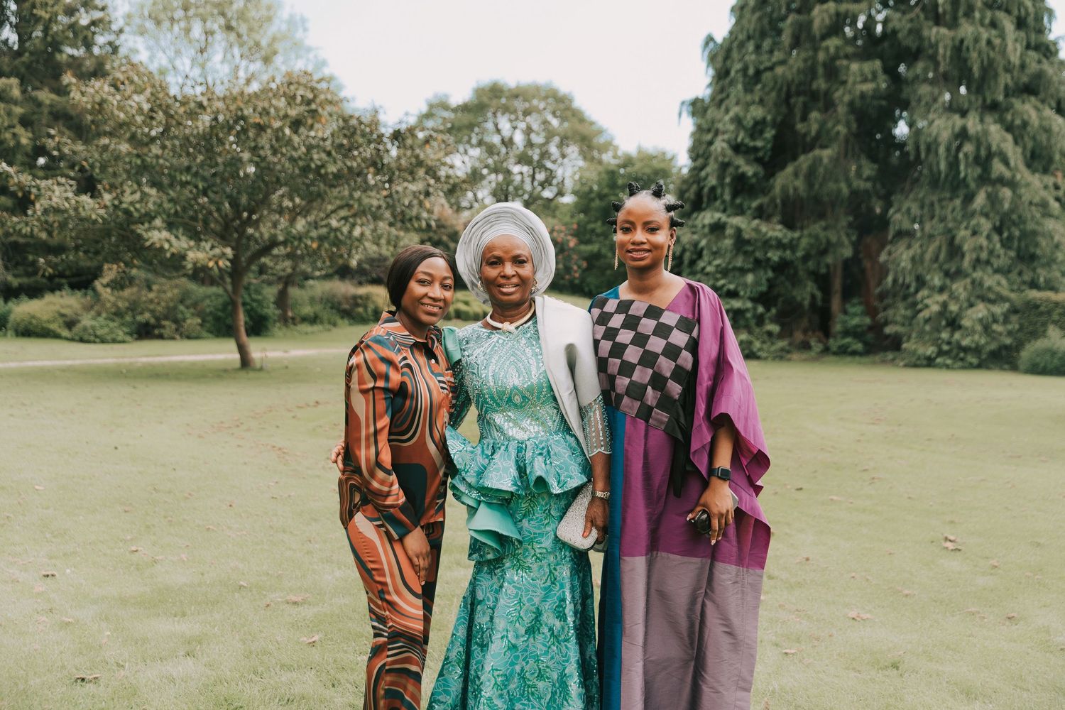 Three people stand together in a grassy outdoor setting wearing colorful clothing and smiling.