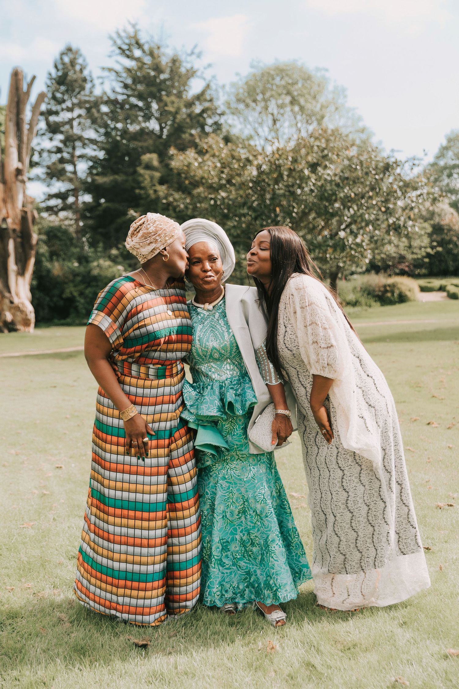 Three women share a joyful moment outdoors wearing colorful dresses including plaid, teal, and white lace patterns on a grassy lawn.
