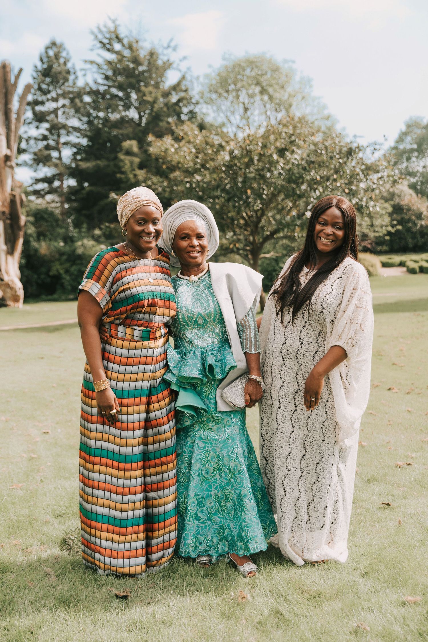 Three people in colorful dresses share laughter and joy while standing together on a grassy lawn in a park setting.