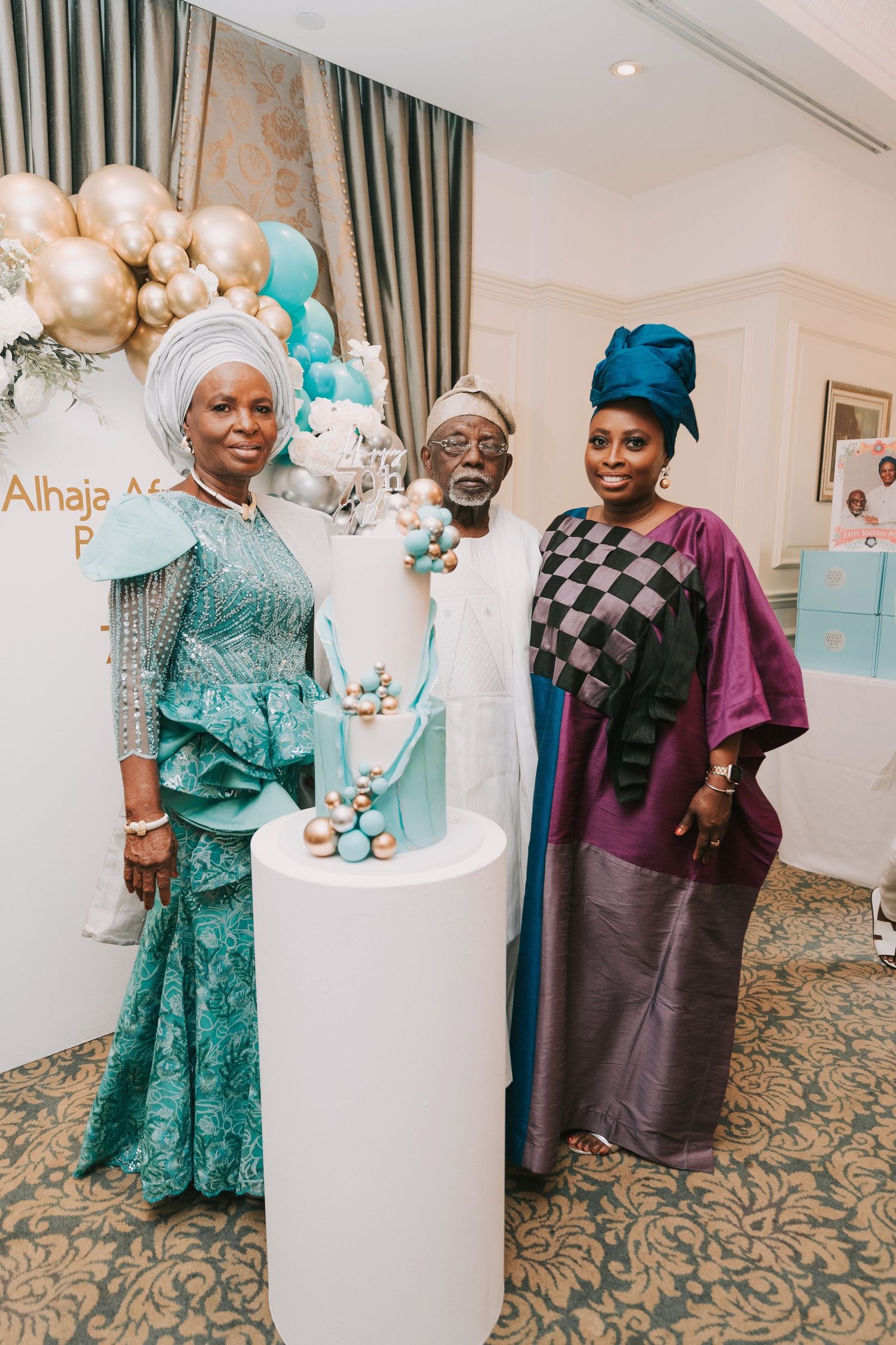 Guests in elegant African formal wear pose by a decorated cake display with balloons and teal accents.