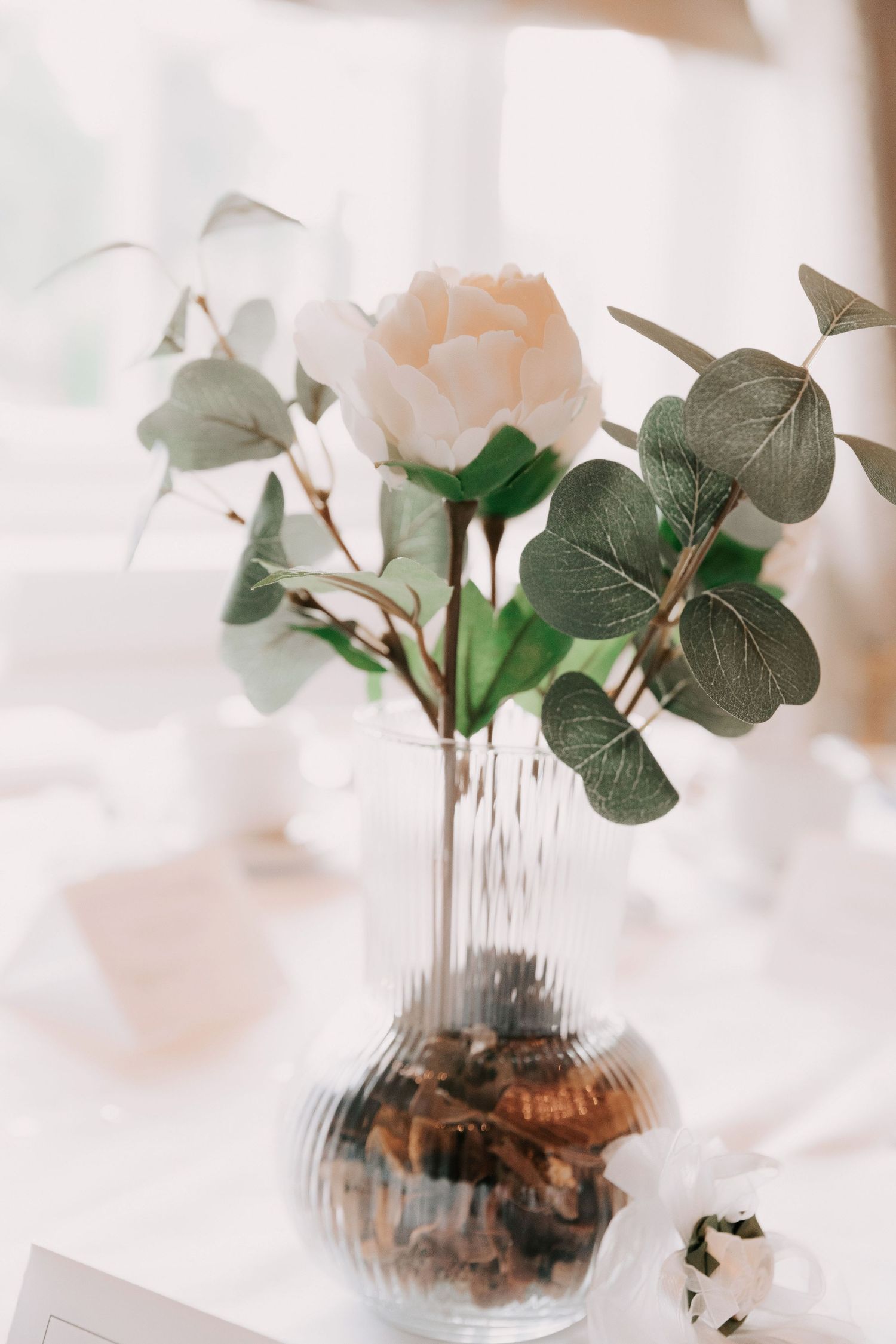 A delicate pink rose and eucalyptus leaves arranged in a clear glass vase with decorative stones at the bottom.