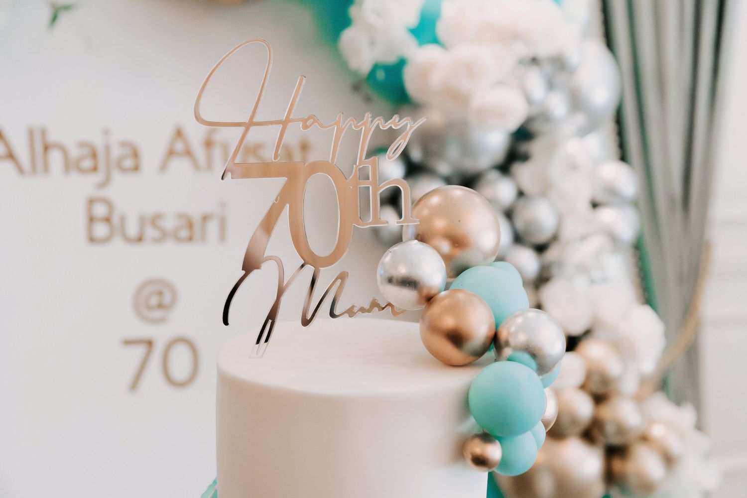 Elegant white wedding cake decorated with turquoise and gold balloon garland and gold lettering for Alhaja at 70th celebration.