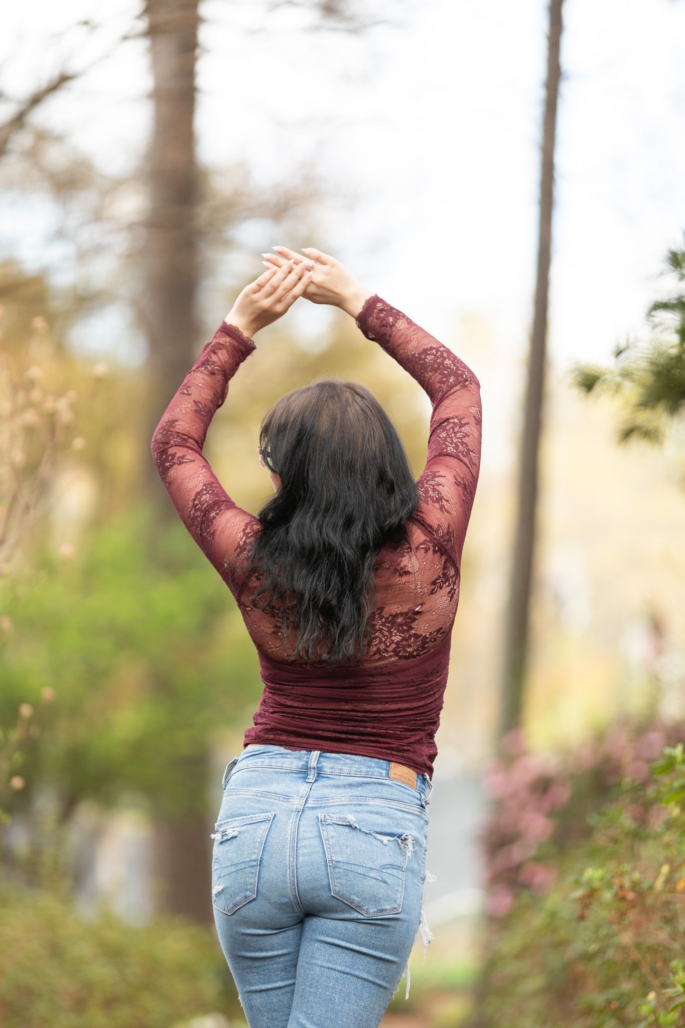 Young woman in ripped jeans and burgundy lace top stands arms crossed on a garden path surrounded by blooming flowers.