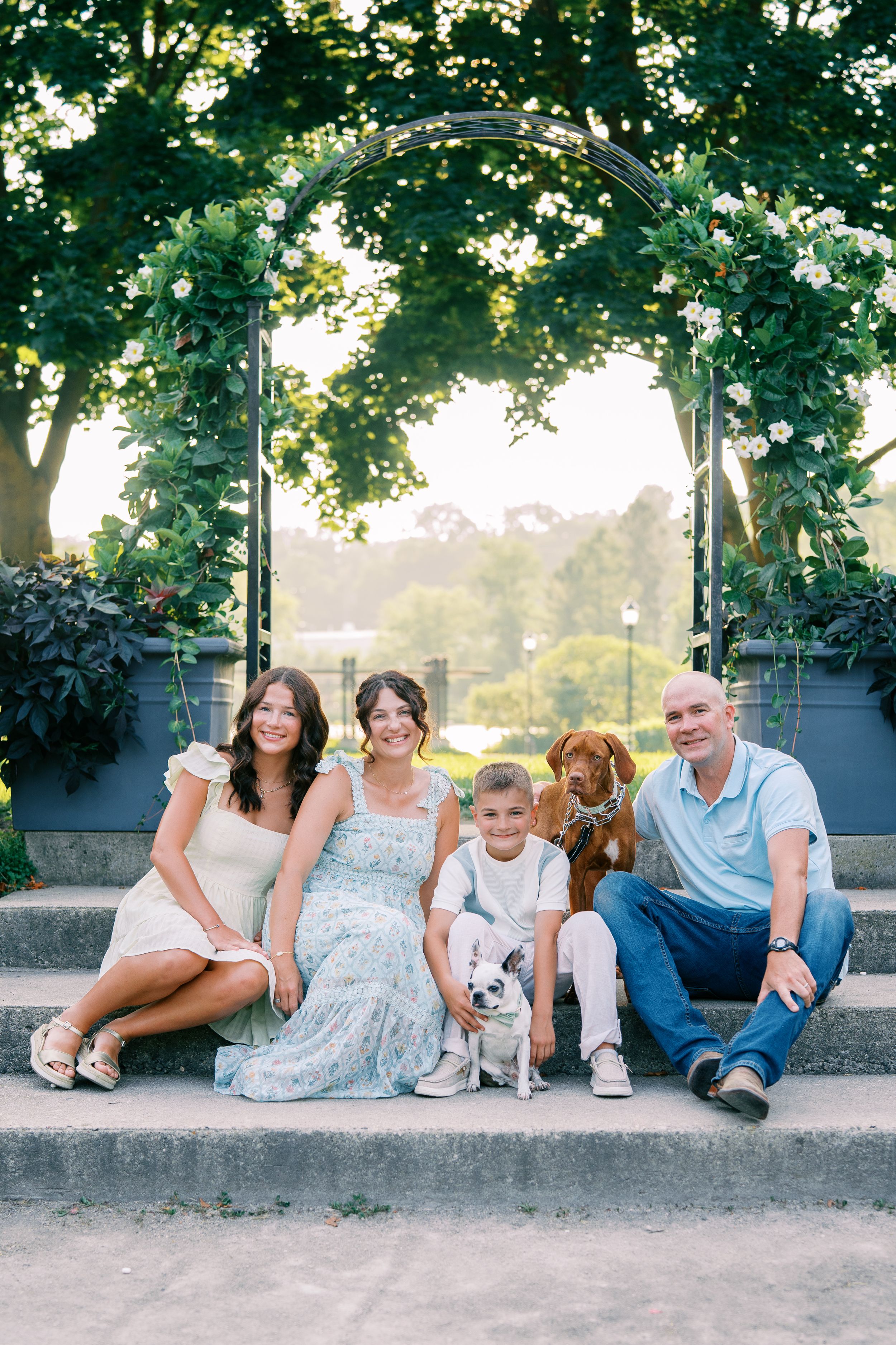 A family with their dogs sits together under a floral archway in a garden setting.