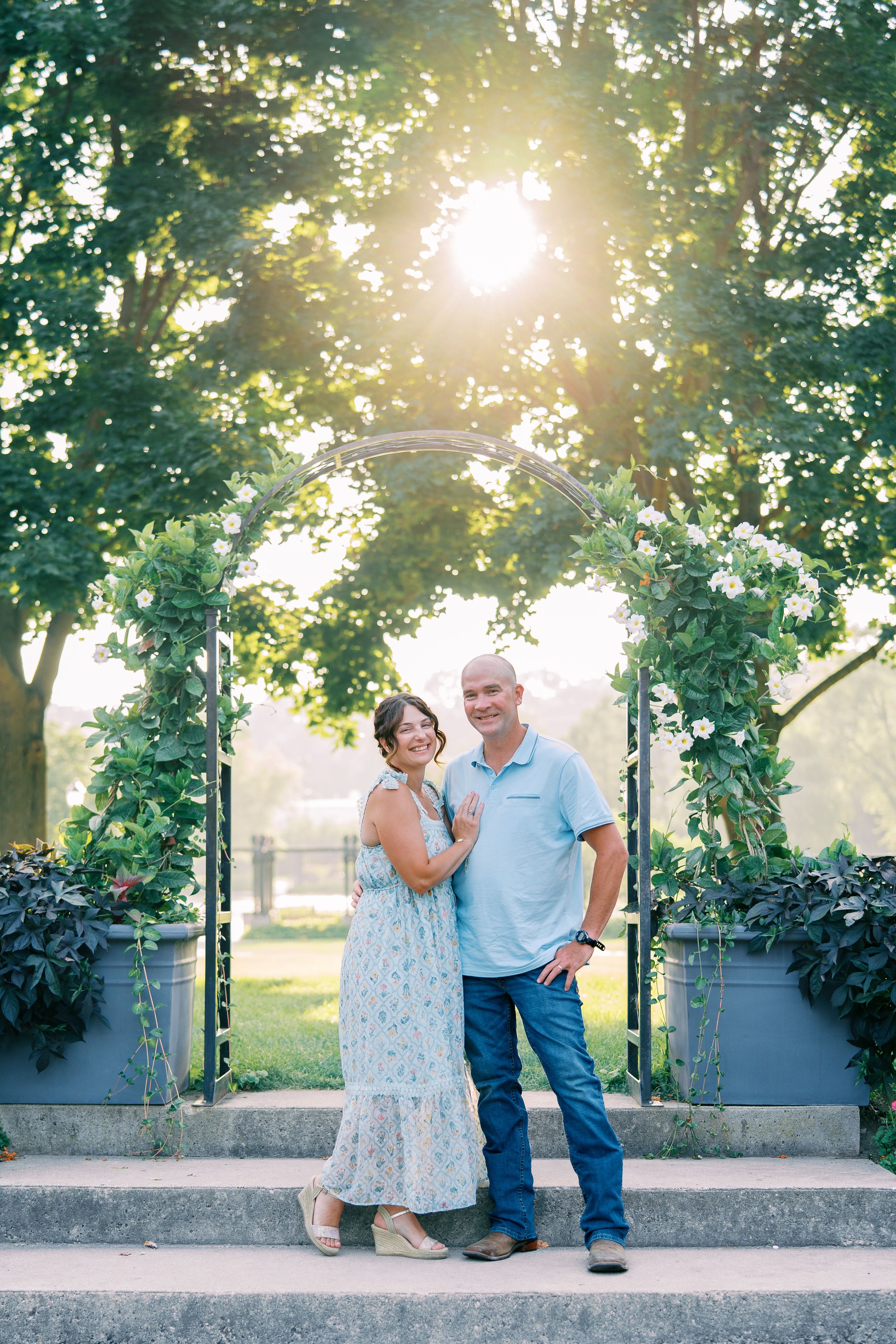A couple stands together under a sunlit garden arch with greenery and flowers on a summer evening.