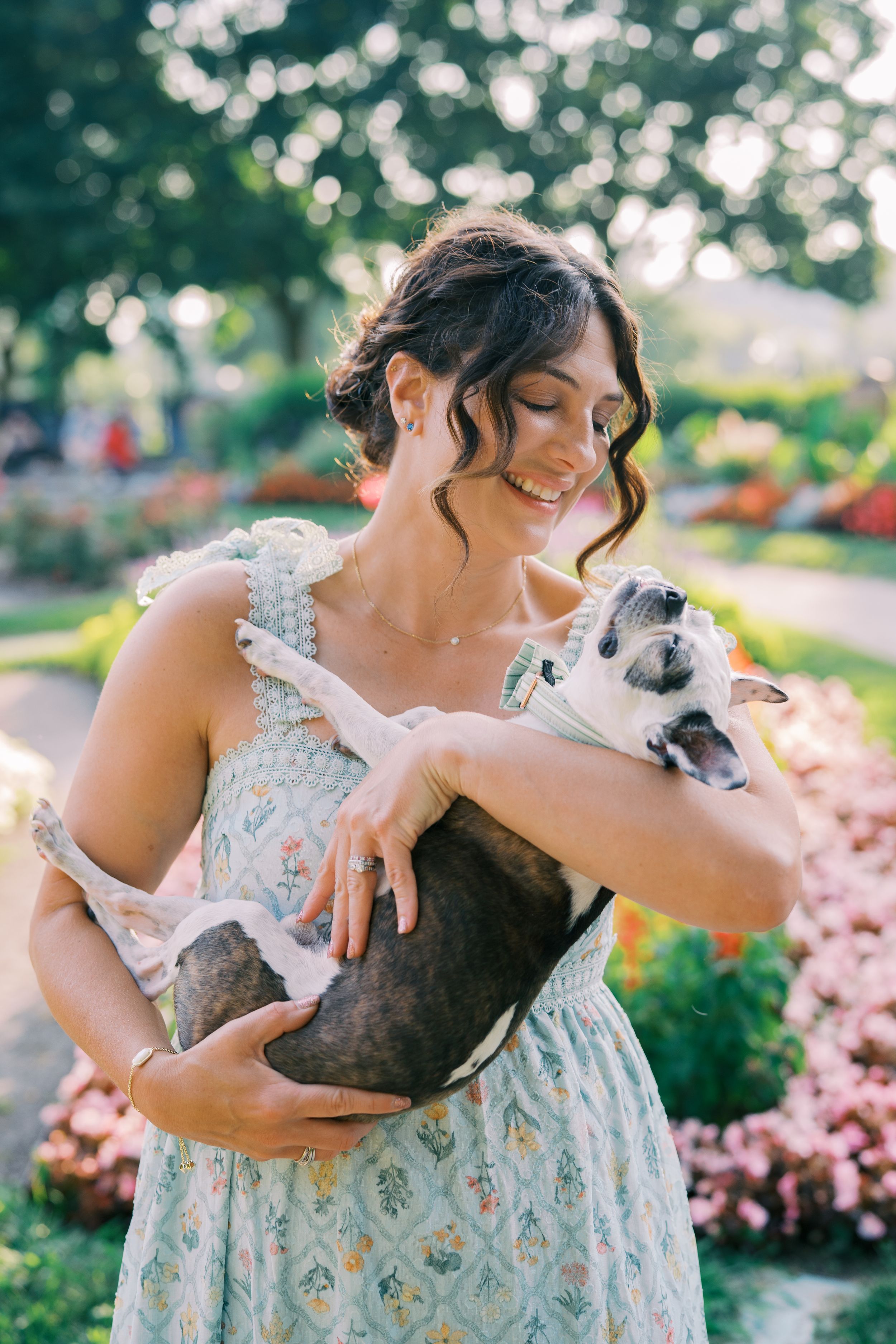 Woman in floral dress cuddles and plays with small dog in sunny garden setting.
