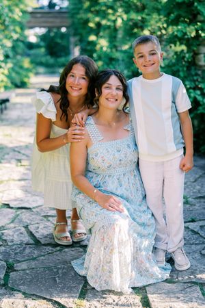 A family of three poses outdoors on a stone path surrounded by greenery on a sunny summer day.