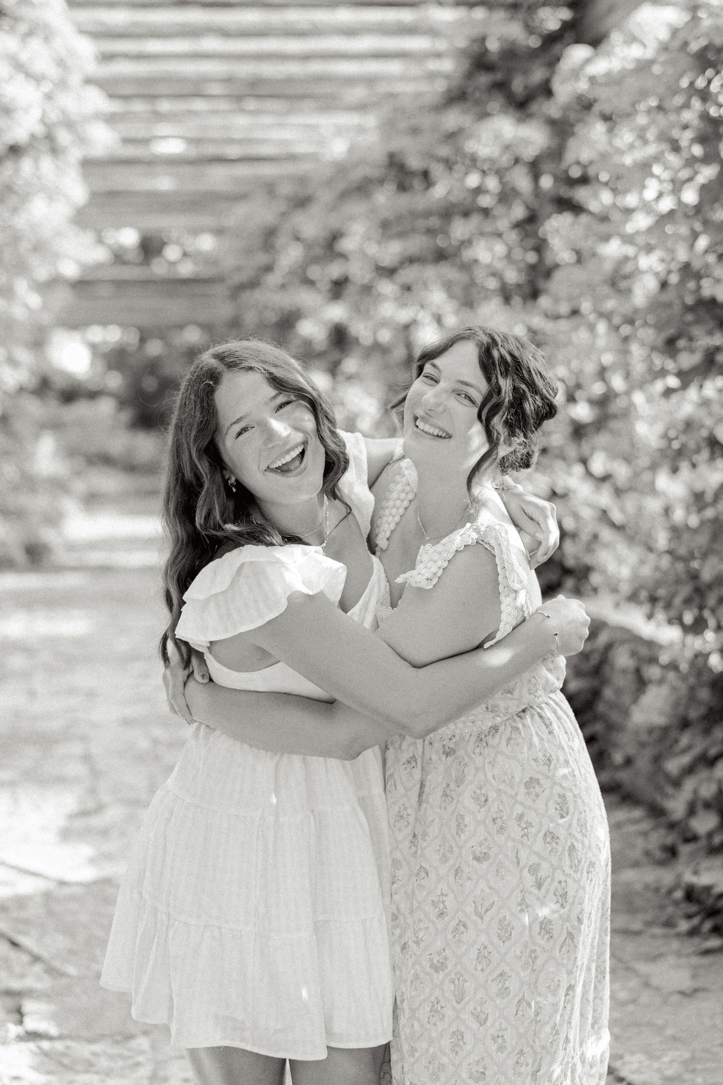 Two people in white dresses share joyful embraces in a sunny garden setting with steps in the background.