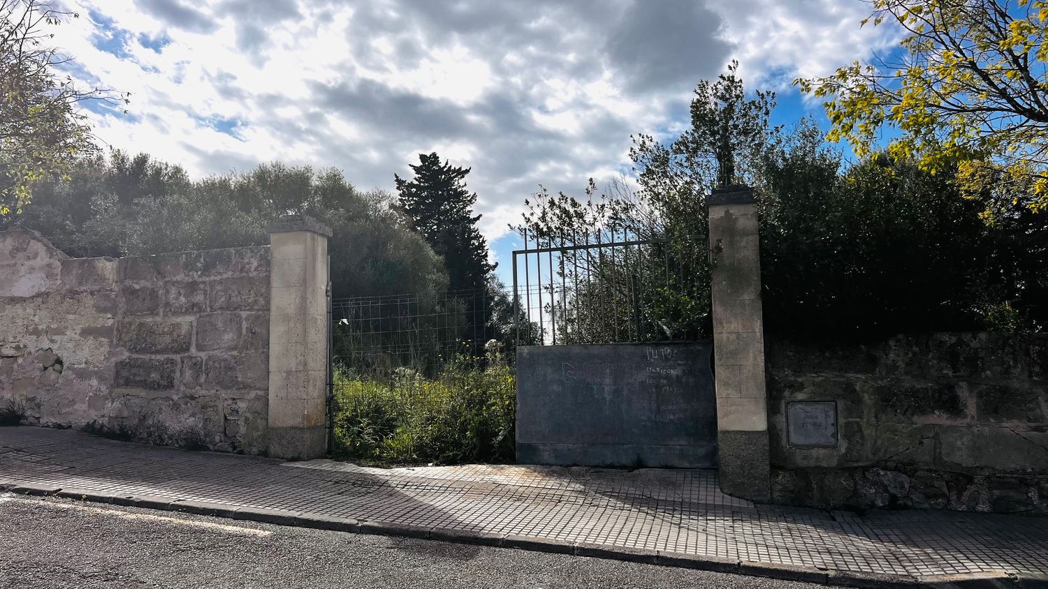 An ornate metal gate stands between two stone pillars in a grassy area with trees.