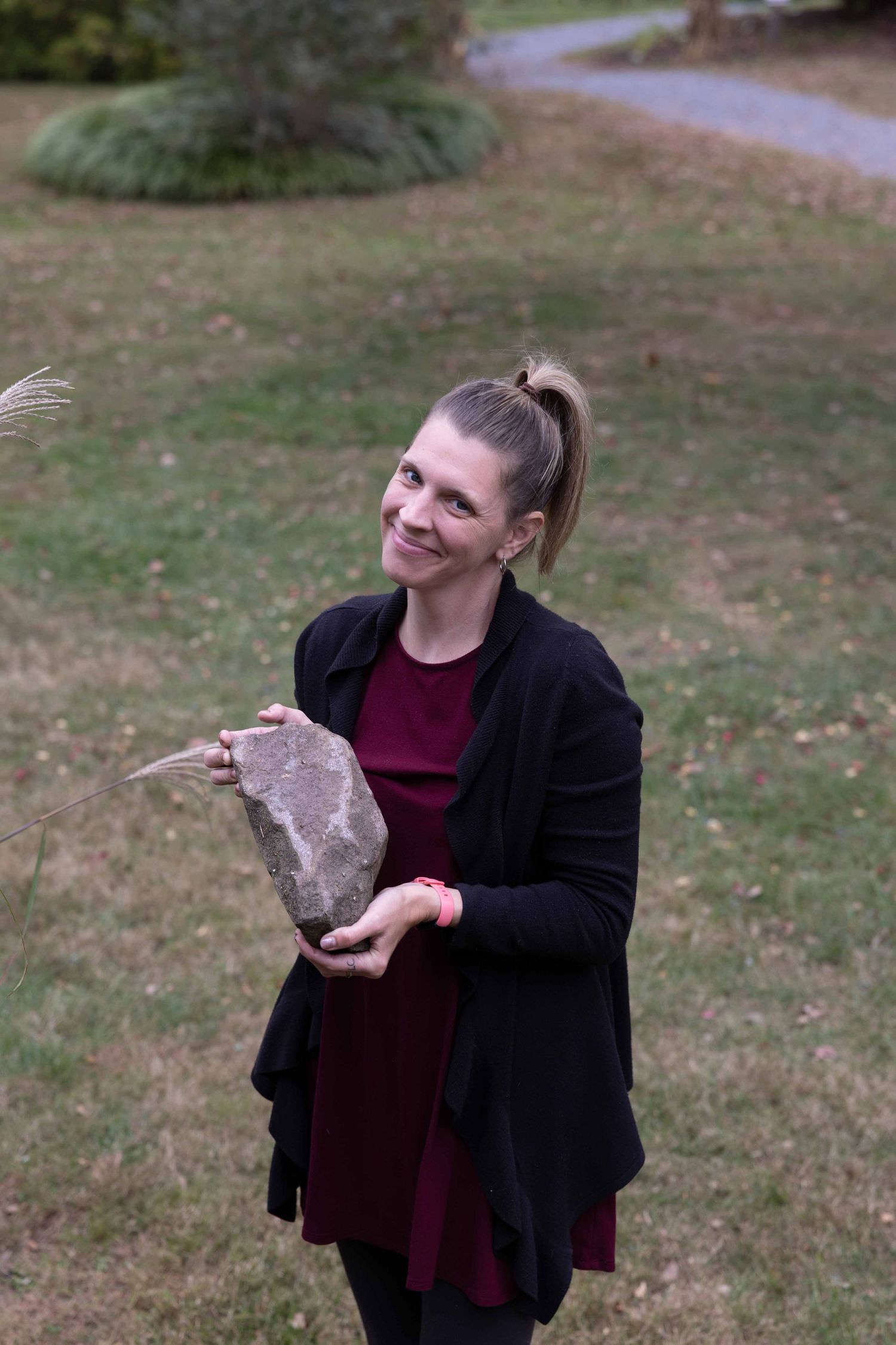 A person in a black blazer and burgundy top poses playfully outdoors on a grassy lawn.