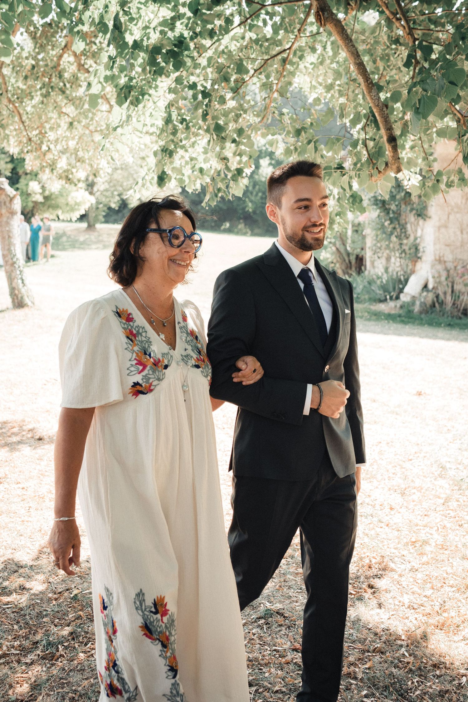 Wedding couple walking through a sunlit garden path with trees overhead.
