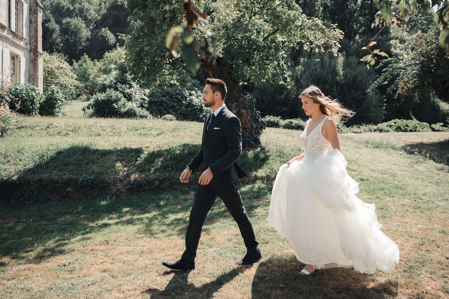 A bride in a flowing white gown and groom in dark suit walk together on sunny grass lawn near trees.
