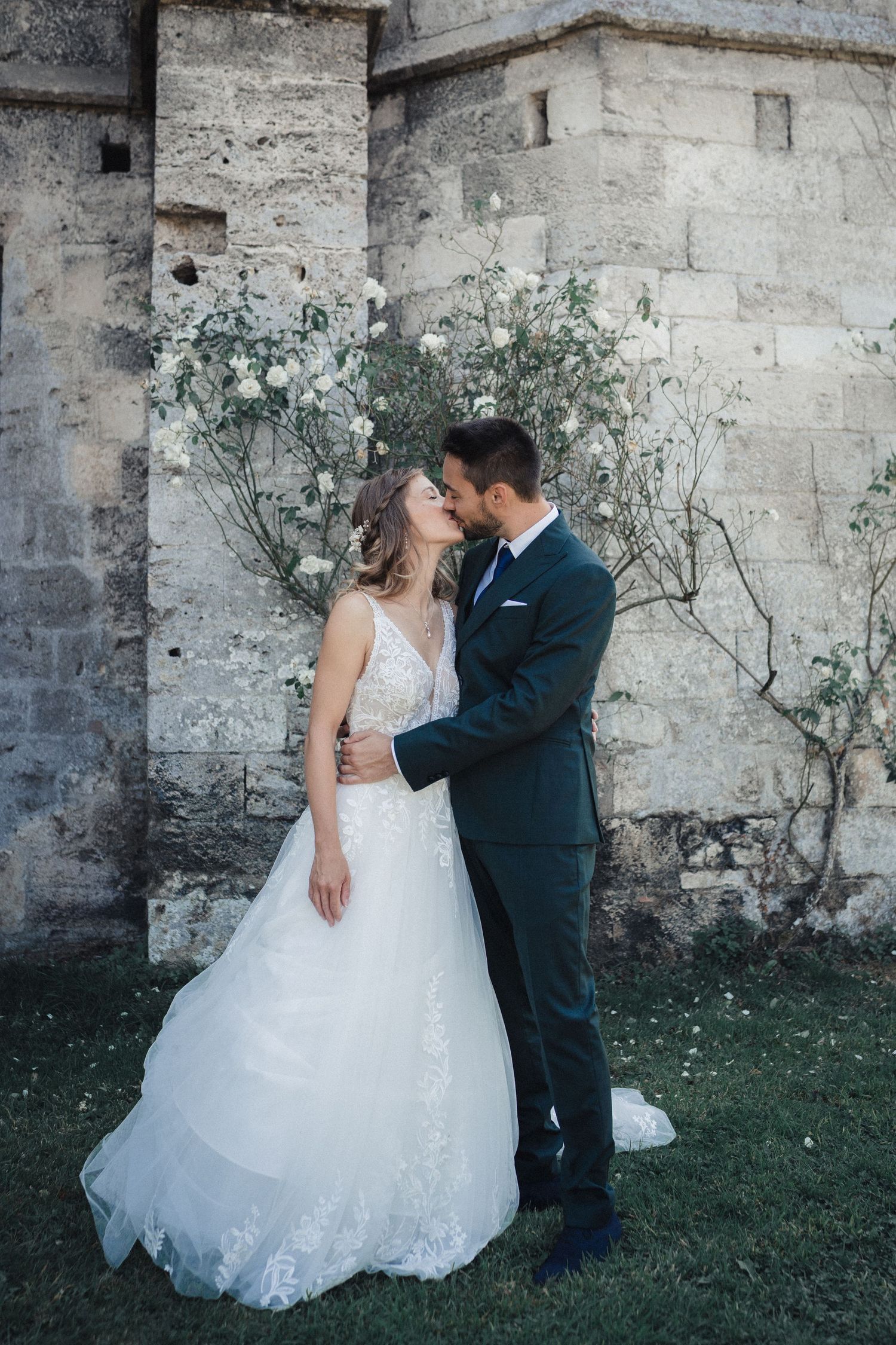 A romantic wedding couple embraces against a stone wall with delicate white flowers in an elegant outdoor setting.