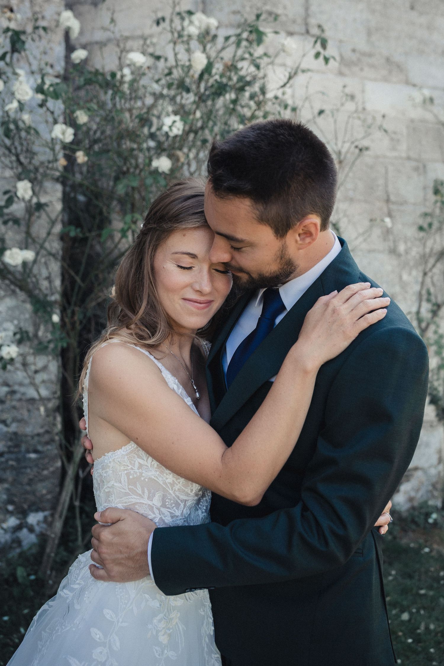 Romantic wedding couple embraces in front of stone wall with climbing vines.