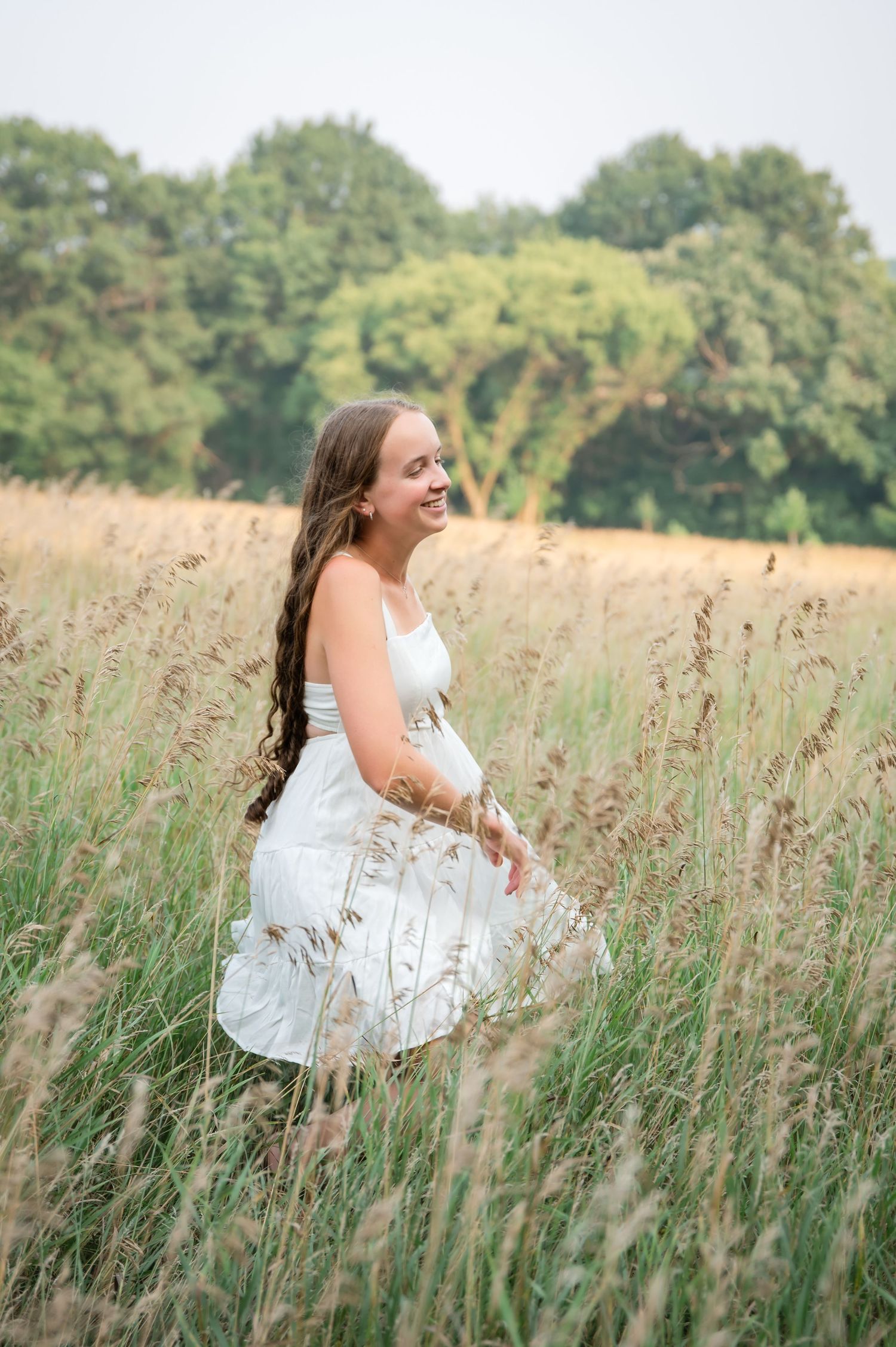 A woman in a white dress laughs joyfully while sitting in a field of tall summer grass with trees in the background.