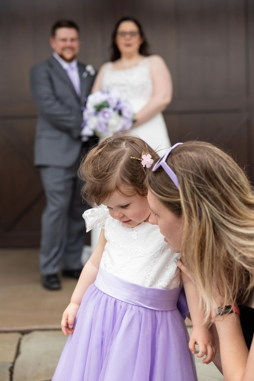 A young flower girl in a white and purple dress looks down while standing with others at a wedding ceremony.