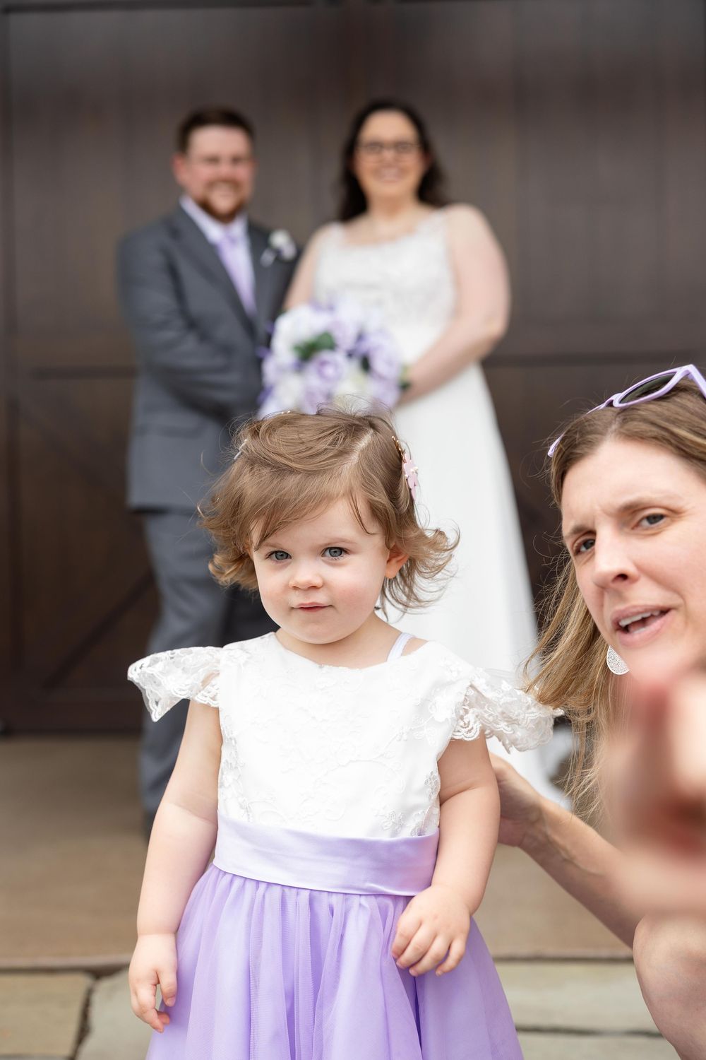A small child in a white and purple dress stands in front of a wedding couple at an outdoor ceremony.