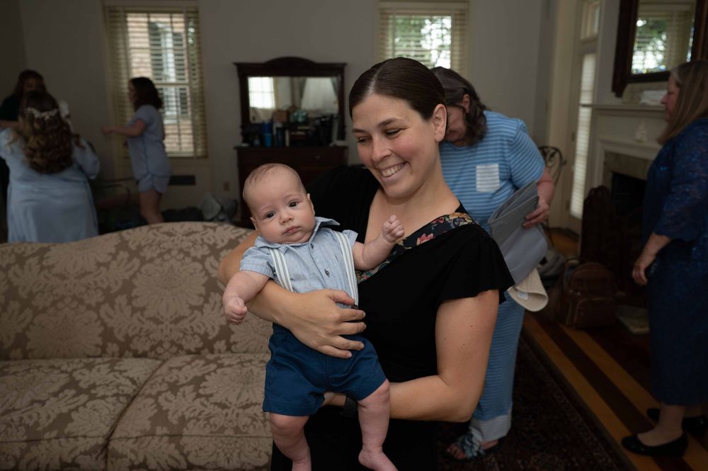 A woman in a black top holds a baby wearing blue in a living room setting with others visible in the background.
