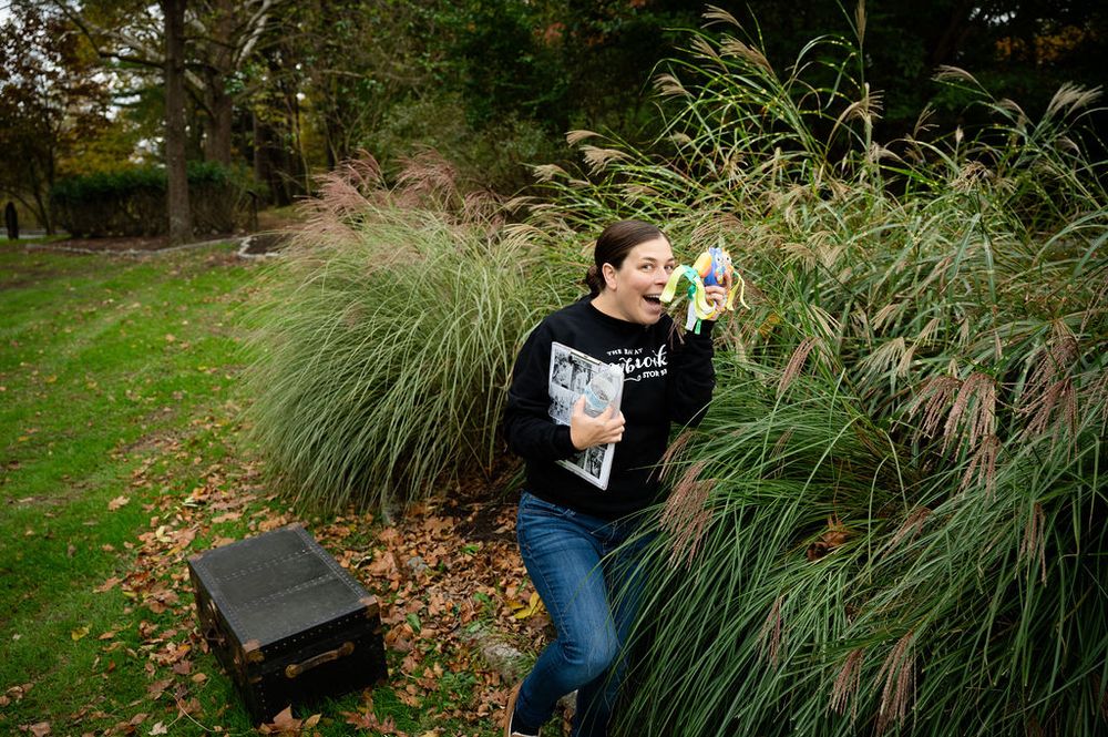 Someone in a black sweatshirt sits on a bench near tall ornamental grasses in an autumn garden setting.