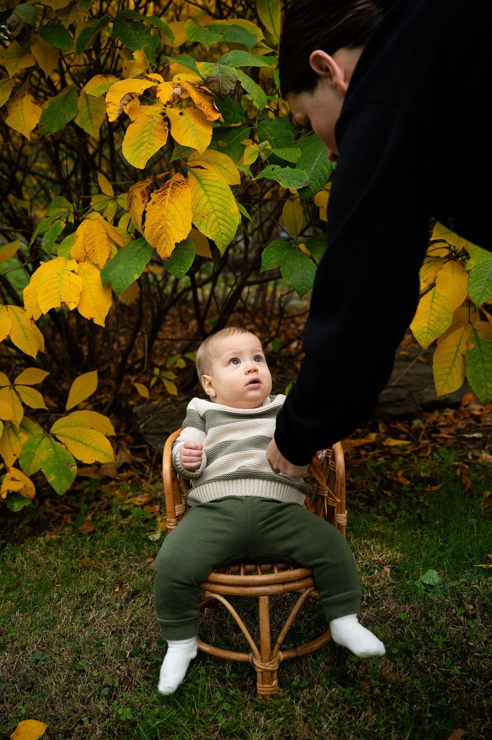 A small child sits in a rattan chair surrounded by autumn leaves with yellow foliage in the background.
