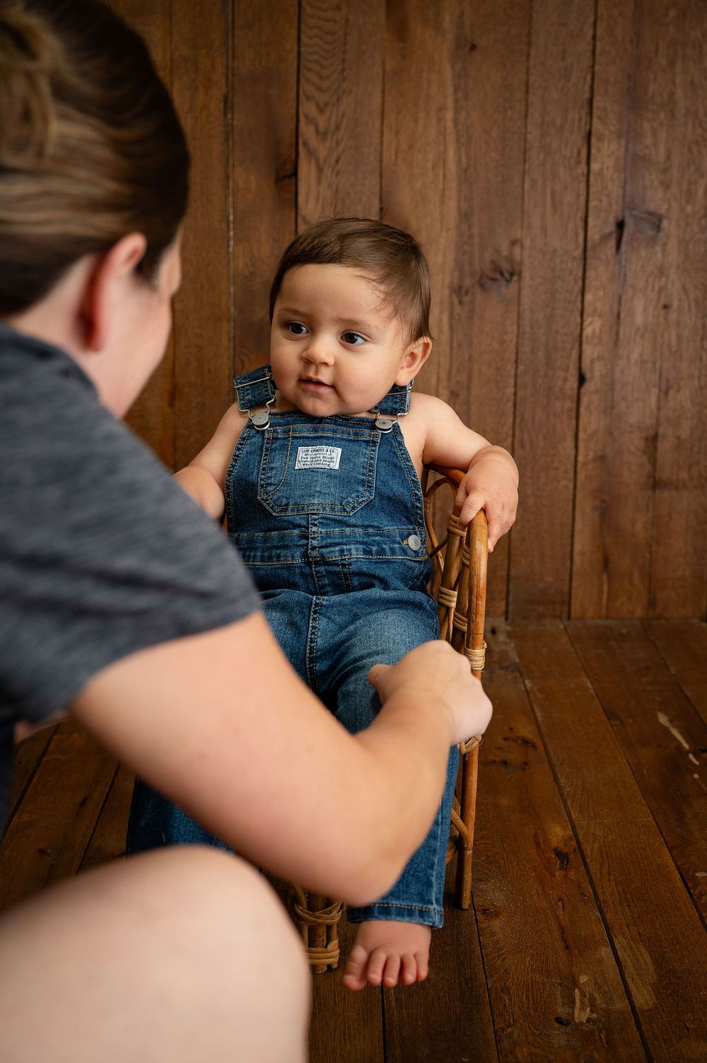 A baby in denim overalls sits in a wooden chair against a rustic wood panel wall.