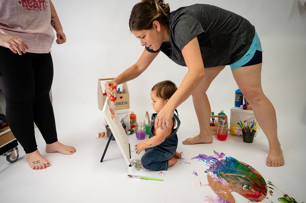 A young child sits on the floor with art supplies while an adult assists during a creative painting session.