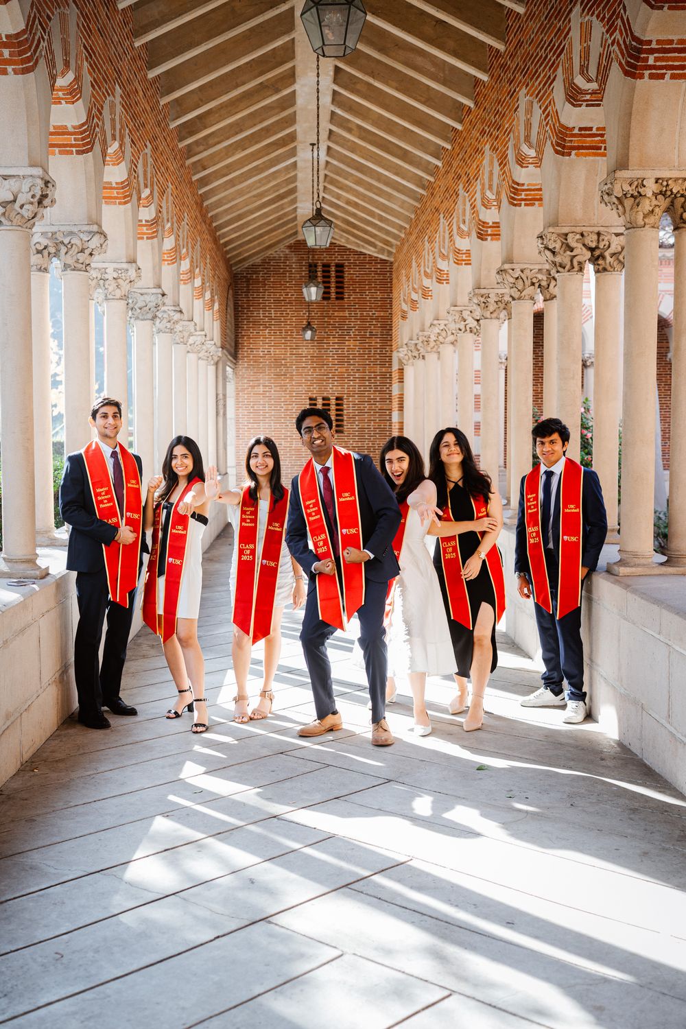 Graduates in black attire with red stoles pose creatively in a sunlit USC Mudd Hall columned hallway for a panoramic photo series.