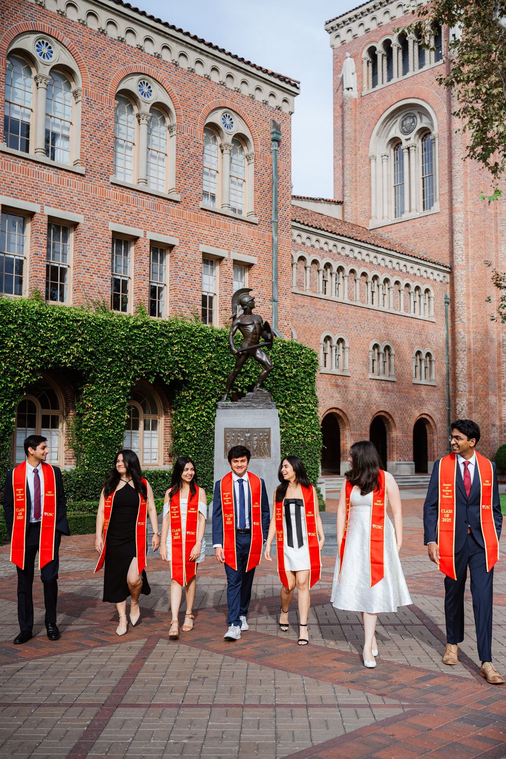 Graduates in academic regalia gather around a statue at USC's brick Romanesque Revival campus building.