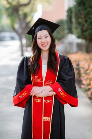 Graduate in black and red academic regalia with USC stole and graduation cap smiles on campus walkway.