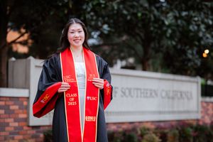 USC graduate in red and black academic regalia poses outdoors for graduation photos near brick buildings.