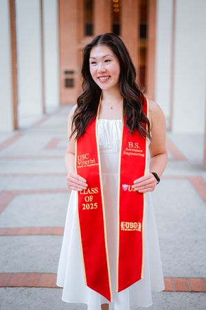 USC graduate wearing white dress and red graduation stole poses for photos in outdoor hallway setting.