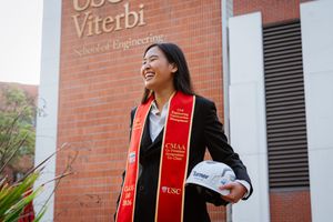 USC Viterbi School of Engineering graduate smiles holding a hard hat, wearing a red graduation stole outdoors.