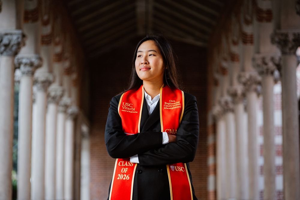 Young woman in black jacket and red USC Class of 2026 graduation sash stands confidently in an archway.