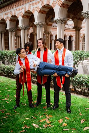 Four USC graduates in red sashes playfully lift a friend on a green lawn.
