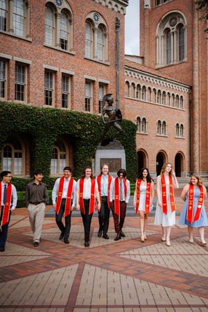USC graduates in red sashes walk together in front of Tommy Trojan statue on campus.