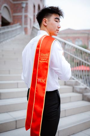 Graduate in formal attire wearing a red and gold Viterbi USC graduation stole poses on stone steps.