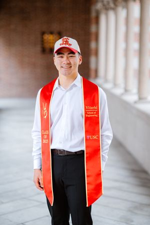 Graduate wearing red USC graduation stole and red baseball cap poses in columned hallway.