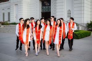 USC graduates wearing red stoles and black gowns form a line against the white exterior wall of Griffith Observatory.