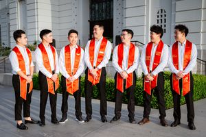 A row of USC graduates in black gowns and red stoles pose together outside a building.