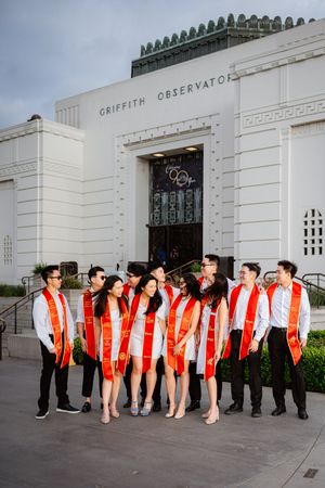 A line of USC graduates in red stoles pose together in front of the white facade of Griffith Observatory.