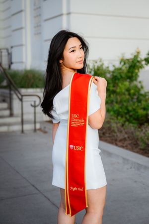 A graduate in white attire and red USC graduation stole poses for portraits on campus.