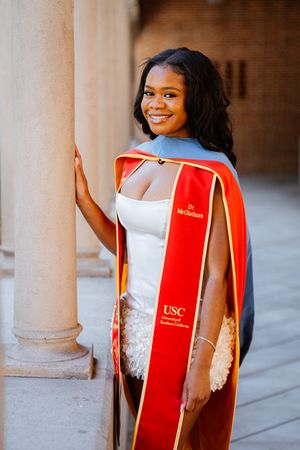 A smiling graduate poses in a red USC University of Southern California academic regalia gown beside a stone column.
